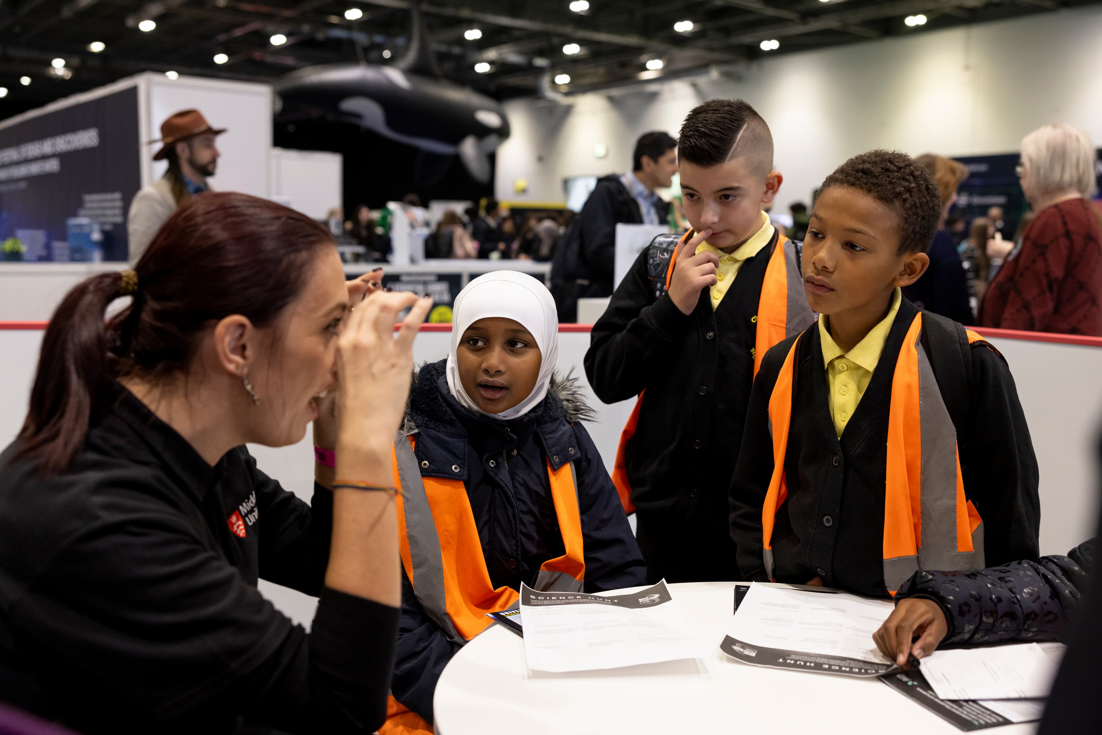 A group of children wearing orange safety vests listens attentively to a woman explaining something at a table during an educational event. The background shows a busy indoor setting with exhibits and a large inflatable orca.