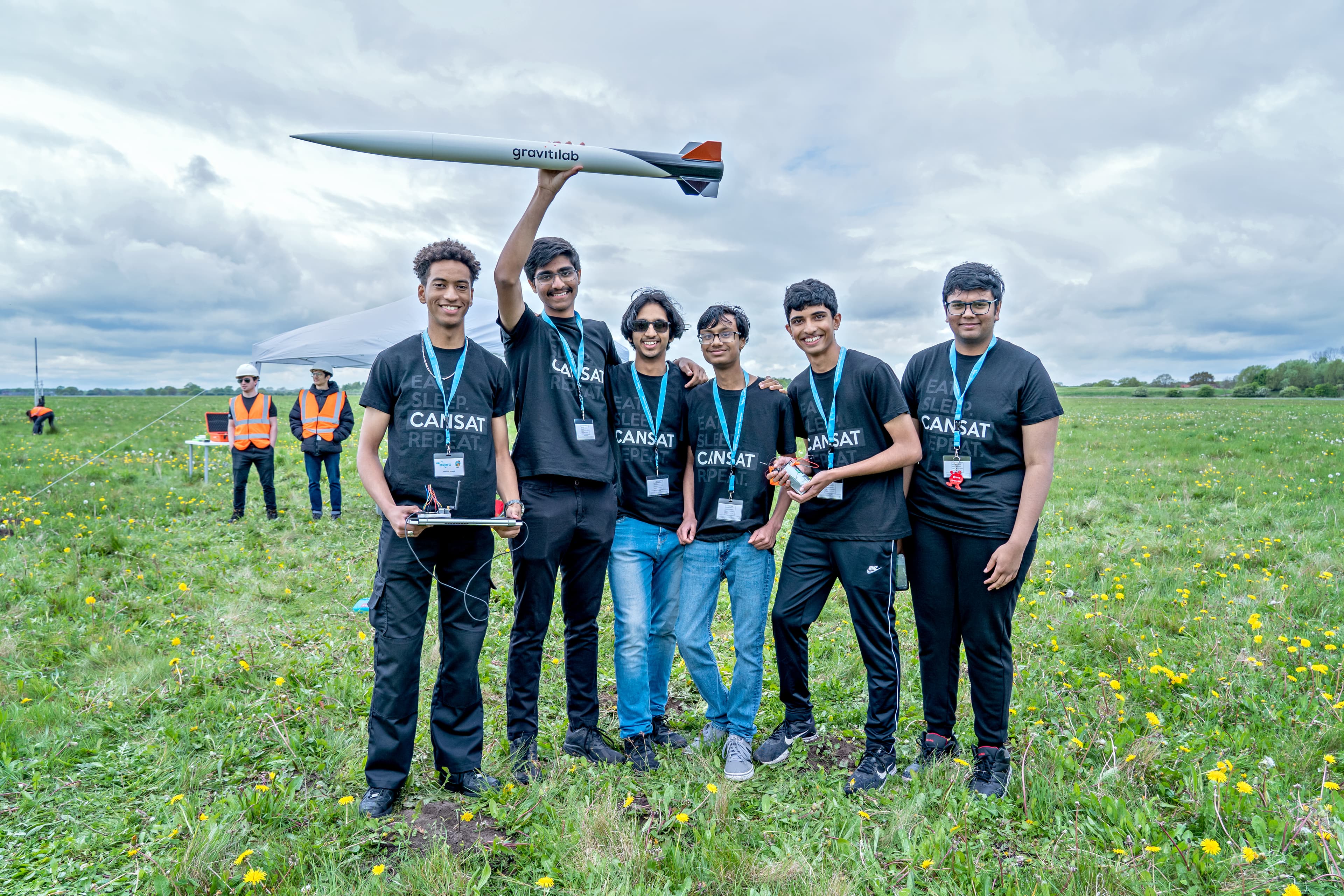 Group of students taking part in CanSat competition, stood on grassy field and raising a rocket in the air.